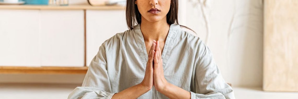 Woman meditating with eyes closed, hands together in peaceful indoor setting.